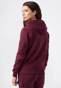 Sudadera burdeos con capucha ajustable, puños acanalados y una textura suave. Combinada con pantalones a juego, mostrando un diseño de outfit casual.