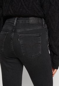 Μαύρες τζιν παντελόνες με στενή γραμμή, που διαθέτουν κλασικό σχέδιο πίσω τσέπης και ετικέτα στη σκοτεινή ζώνη. Η υφή είναι λεία και ελαφρώς φθαρμένη.