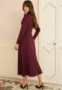 Rochie bordeaux cu mânecă lungă, cu un corset ajustat, fustă amplă și un accent negru la talie, asociată cu pantofi cu toc înalt negri. Fundal deschis.