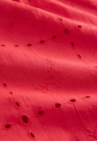Rød stof med blomsterbroderi og perforerede detaljer, der har en glat tekstur og synlige sy- og broderingsmønstre.