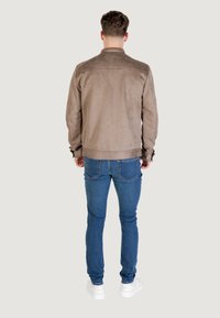 Giacca di suede marrone con una texture liscia, colletto chiuso e polsini a coste, abbinata a jeans blu e sneakers bianche. Vista da dietro.