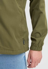 Jachetă verde măsliniu cu manșete și tiv elastic. Fabricată dintr-un material neted, ușor. Prezentă o etichetă neagră cu detaliul "BLEND".
