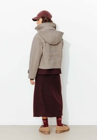 Veste à capuche grise, robe en tricot bordeaux côtelée, bottes marron et bonnet bordeaux. Textures douces et look superposé avec des détails de couture visibles.