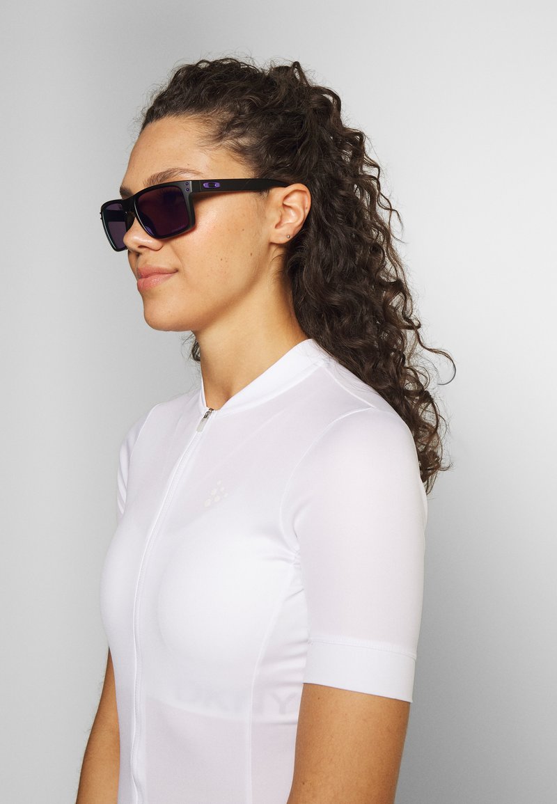 Schwarze rechteckige Sonnenbrille mit dunklen Gläsern, die einen leichten Farbverlauf aufweisen. Vom Träger getragen, der ein enges, kurzärmliges weißes Radtrikot anhat.