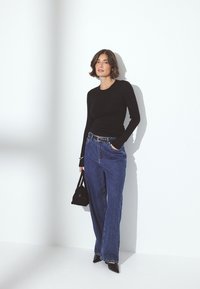 Čierny top s dlhými rukávmi, modré vysoké jeansy, čierna kabelka, špicaté čierne ihličky. Hladká látka, voľný strih, jednoduchý dizajn na bielom pozadí.