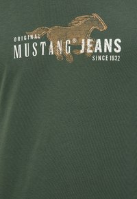 Green cotton t-shirt featuring a brown horse graphic and white text reading "Original Mustang Jeans Since 1932." Smooth texture.