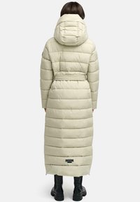 Lange beige pufferjas met een capuchon, voorzien van doorgestikte delen, een ceintuur in de taille en een logopatch aan de zoom. Draagt het met zwarte laarzen.