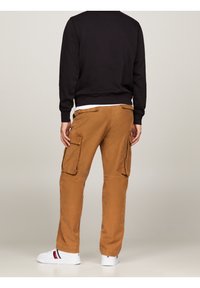 Braune Cargo-Hosen mit Seitentaschen, lockerem Schnitt und Kontrastnähten, kombiniert mit einem schwarzen Langarmshirt und weißen Turnschuhen.