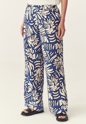 Luźne niebieskie spodnie w białe tropikalne liście noszone z czarnymi sandałami na odkryte palce i białym topem.