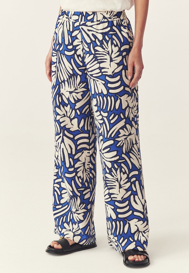 Pantalon ample bleu avec imprimé feuilles tropicales blanches, porté avec des sandales noires à bouts ouverts et un haut blanc.