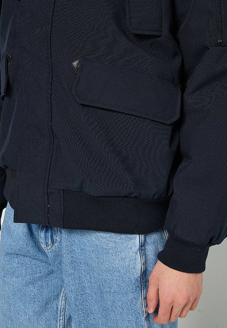 Veste bombardier bleu marine avec une finition texturée, des poignets côtelés et plusieurs poches à rabat. Comprend une fermeture éclair à l'avant et une taille ajustée.