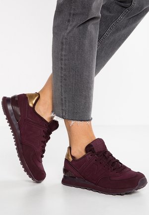 Personne portant un jean gris foncé à ourlet effiloché et des baskets bordeaux foncé avec des accents métalliques dorés au talon sur un sol blanc.