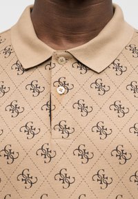 Polo beige avec deux boutons et un motif géométrique sombre répétitif présentant des formes imbriquées sur le tissu.