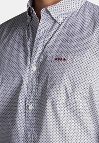 Overhemd met knoopjes in witte stof met een geometrisch blauw en zwart ruitpatroon. Bevat een puntige kraag en een klein rood logo-accent.
