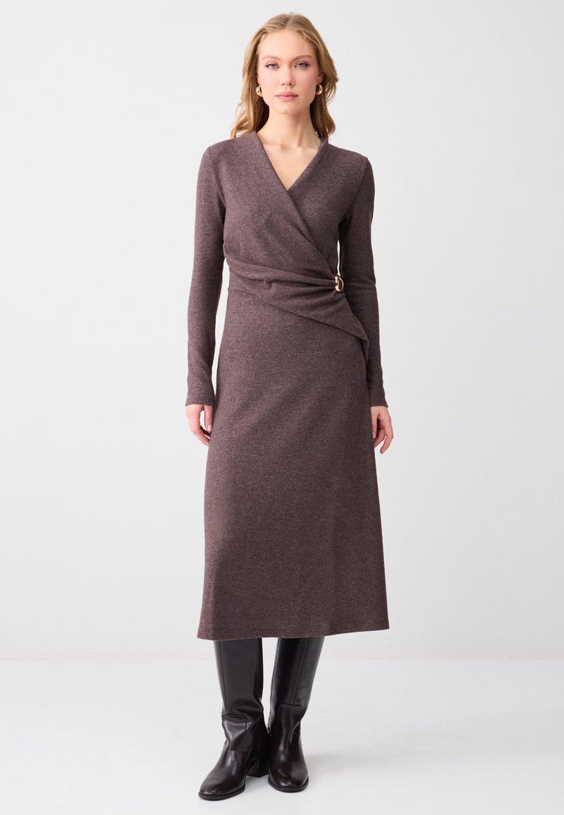 Femme portant une robe midi violette à manches longues et à style portefeuille avec une boucle dorée, associée à des bottes noires montantes, debout devant un fond uni.