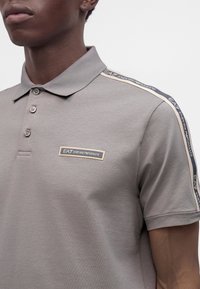 Νέος φοράει γκρι πόλο EA7 Emporio Armani με λογότυπο στο στήθος και ταινία με λογότυπο στους ώμους.