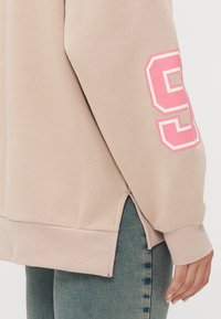 Felpa beige con una grafica rosa "9" sul manico sinistro, polsini a costine e fessure laterali all'orlo per facilitare i movimenti.