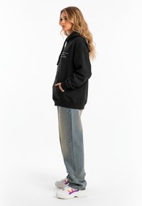 Felpa nera con tasca frontale, cappuccio regolabile con coulisse e dettagli in bianco. Indossata con jeans azzurri chiari e sneakers multicolori.