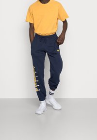 Gul bomullst-shirt kombinerad med marinblå mjukisbyxor med vit och gul "Adidas"-text längs sidan och en liten logo på höften.