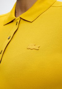 Polo jaune avec col et boutons, arborant un logo crocodile texturé brodé sur la poitrine.