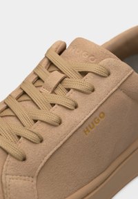 Μπεζ πάνινο παπούτσι σουέντ με κορδόνια ίδιου χρώματος, χρυσό λογότυπο «HUGO» στο πλάι και επενδυμένο γιακά σε ουδέτερο φόντο.