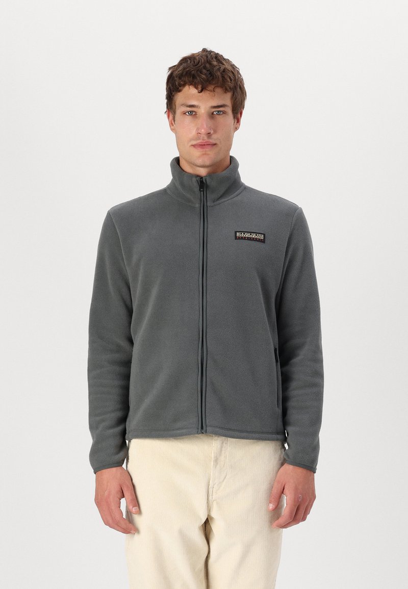 Giacca in pile grigia con colletto alto, zip completa e un patch rettangolare sul petto sinistro, abbinata a pantaloni di velluto a coste beige.