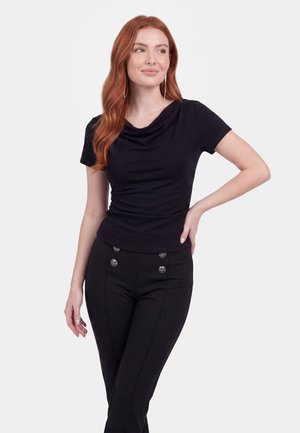 Donna con lunghi capelli rossi che indossa una maglietta nera a maniche corte e pantaloni neri con bottoni decorativi, in piedi con la mano sul fianco.