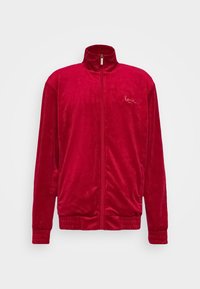 Veste zippée en velours rouge avec un col montant, des poignets et une taille élastiques. Présente un logo brodé discret sur la poitrine. Texture lisse.