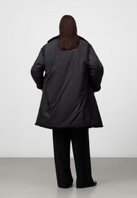 Person som står med ryggen mot kameran, iklädd en svart överdimensionerad vinterjacka med pälsdetaljer och lösa svarta byxor, mot en neutral ljus bakgrund.