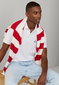 Jonge man die lichtblauwe spijkerbroek, wit poloshirt en een rood-wit gestreepte trui over zijn schouders draagt, zittend op een houten bank.