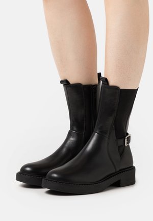 Svarta ankellånga stövlar i slätt läder med dragkedja på sidan, elastiska paneler och en metallspänndetalj på utsidan. Klumpig gummisula.