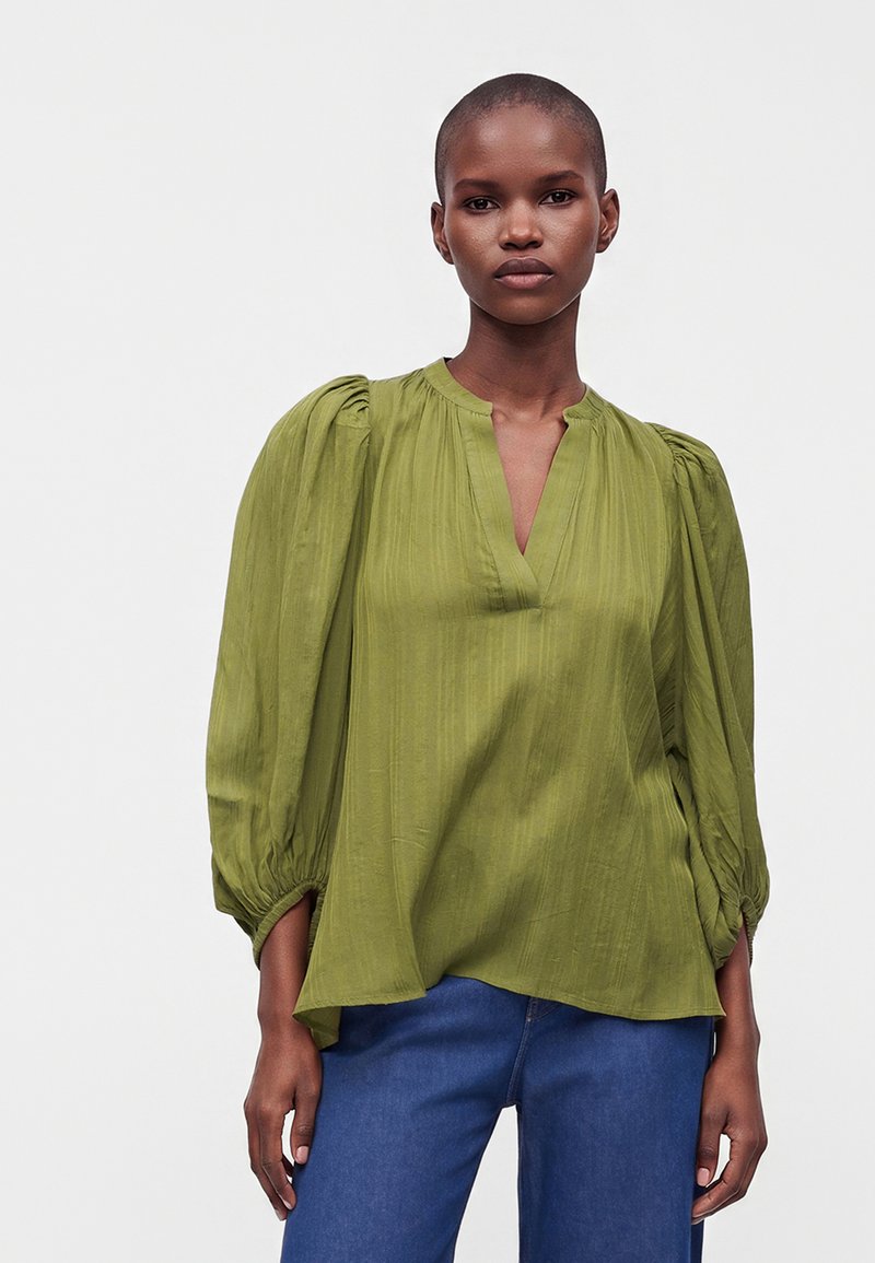 Femme aux cheveux courts portant un chemisier ample vert olive avec des manches bouffantes froncées et un jean bleu sur un fond uni.