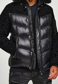 Sort dunjakke med en blank finish, der har et mønstret stof på skuldrene, lynlås lukning og pelsbeklædt hætte.