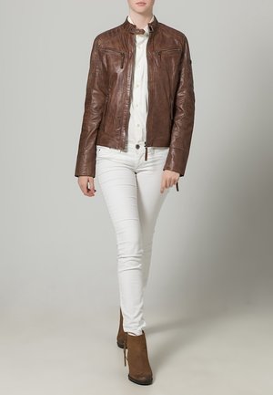 Vrouw die loopt, gekleed in een bruine leren jas, wit overhemd met knopen, witte broek en bruine enkellaarzen, tegen een effen neutrale achtergrond.