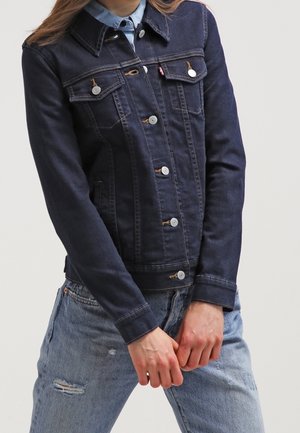 Persona con una chaqueta vaquera ajustada de color azul oscuro con cierres de botones sobre una camisa azul clara, combinada con vaqueros claros desgarrados.