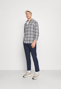 Sjekket, grå og hvit skjorte med rullede ermer, marineblå bukser, beige joggesko og en lys caps. Modellen står mot en ensfarget bakgrunn.
