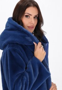 Cappotto in pelliccia sintetica blu con un ampio cappuccio, caratterizzato da una texture morbida e maniche voluminose. Dettaglio con zip visibile sul pannello frontale.