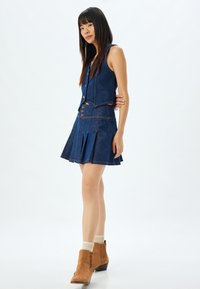 Ensemble composé d'un gilet en denim et d'une jupe bleu foncé. La jupe présente des plis et des détails de boutons. Des bottines beige à la cheville avec des chaussettes crème côtelées viennent compléter le look.