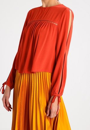 Kobieta w luźnej, czerwonej bluzce z rozcięciami na rękawach i wiązaniami, w połączeniu z plisowaną, pomarańczowo-żółtą spódnicą z wysokim stanem.