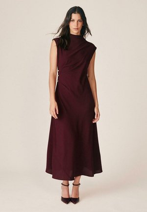 Mujer de pie con un vestido sin mangas, burdeos oscuro, hasta el tobillo con botones laterales, combinado con tacones oscuros con tira en el tobillo.