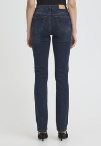 Femme portant un jean droit bleu foncé et des talons noirs, vue de dos sur un fond clair uni.