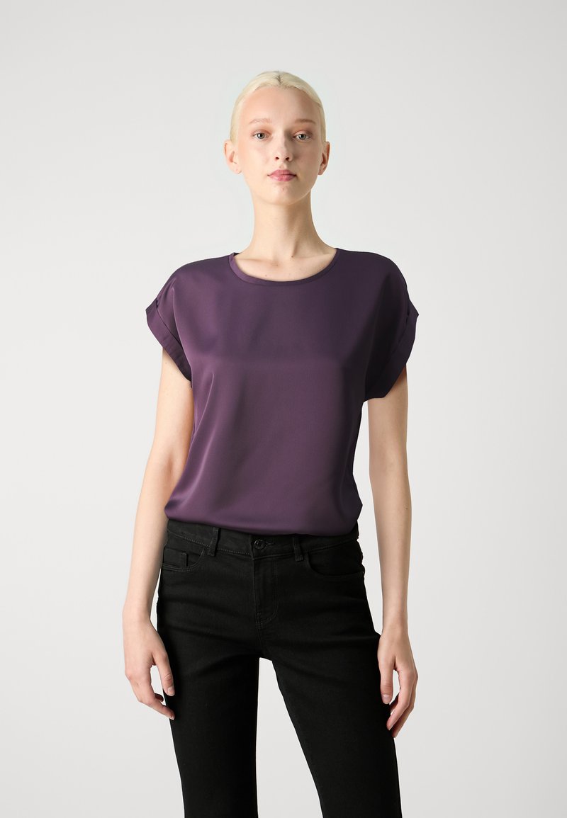 Blusa viola a maniche corte con scollo rotondo e texture liscia, abbinata a jeans neri slim. Design semplice e moderno.