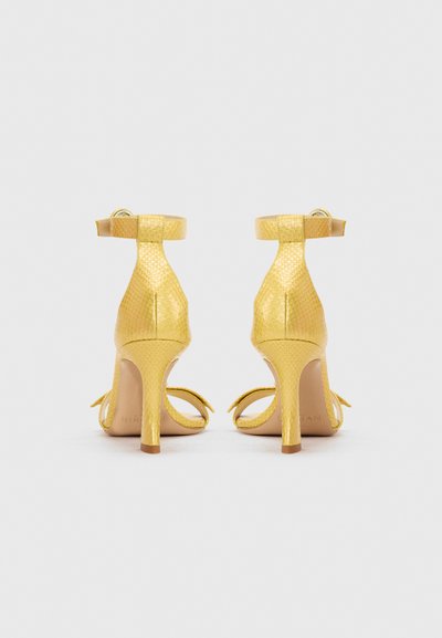 Χρυσά σανδάλια με ψηλό τακούνι και λουράκια στον αστράγαλο με υφή, εμφανιζόμενα από πίσω σε απλό ανοιχτόφωτο φόντο.