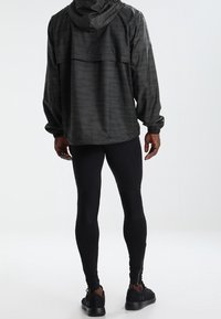 Giacca sportiva nera con cappuccio, caratterizzata da un delicato design testurizzato e polsini elasticizzati, abbinata a leggins neri aderenti e scarpe da ginnastica.