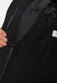 Juodas pūkinis švarkas viduje, rodančiame lygų matinį paviršių su užtrauktuku ir etikete. Medžiaga atrodo lengva ir patvari.