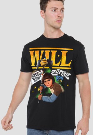Mann trägt schwarzes T-Shirt mit großem gelbem Schriftzug „WILL“ und Cartoon, der eine Axt hält, mit den Texten „Stranger Things Tales from 1985“ und „Zombie Boy“.
