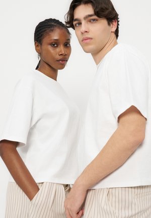 Doi tineri adulți purtând tricouri albe și pantaloni bej cu dungi stând apropiați, privind către cameră, pe un fundal alb simplu.