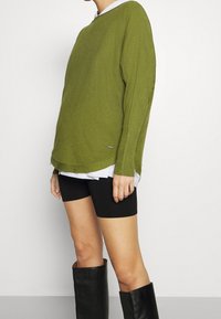 Pull en tricot vert olive à coupe décontractée, à manches longues, avec des fentes sur les côtés, superposé sur une chemise blanche, associé à un short noir et des bottes noires montantes.
