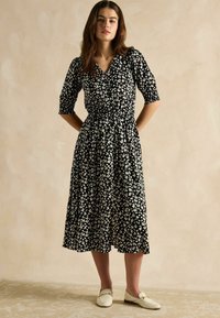 Schwarzes Kleid mit weißem Blumenmuster, V-Ausschnitt, kurzen Puffärmeln, tailliertem Bund und fließendem Rock, getragen mit weißen Slippern.