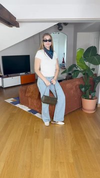 A modell egy fehér pólót, világoskék trapéznadrágot és egy sötétzöld táskát visel, és a barna kordbársony kanapé mellett áll, egy növény társaságában.